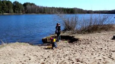 Yetişkinler yakınlarda dinlenirken çocuklar göl kıyısındaki kumda oynarken eğleniyor. Parlak güneş ışığı ve berrak gökyüzü huzurlu göl atmosferini güçlendirir..
