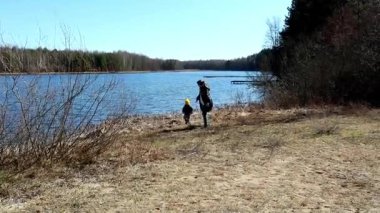 Bir aile, yemyeşil yemyeşil ve parlak mavi gökyüzü ile çevrili sakin bir göl kıyısında gezinir. Yakınlarda bir çocuk oynar, doğada neşeli bir atmosfer yaratır..