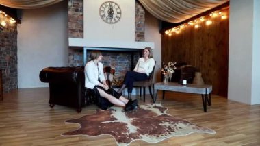 İki kadın, sıcak ve davetkar bir oturma odasında, taş duvarlar ve yumuşak ışıklandırmayla derin bir sohbete girişiyor. Mekanın şık mobilyaları olan rahat bir atmosferi var..