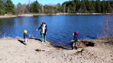 İki çocuk kumlu sahilde oynamaktan zevk alırken bir yetişkin onları dingin bir göl kenarında izliyor. Canlı ayar arka planda berrak mavi su ve ağaçlara sahip.