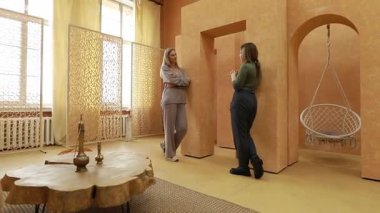 İki kadın yaratıcı bir şekilde tasarlanmış, sıcak tonlarda ve karmaşık dekor elementleriyle dolu bir odada anlamlı bir sohbete girişiyor. Rahatlamış atmosfer açık diyaloğu teşvik ediyor.