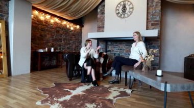 İki kadın zarif bir şekilde tasarlanmış bir salonda rahatça oturup birbirleriyle sohbet ediyor ve eğleniyorlar. Sıcak atmosferde sıcak ışıklandırma ve şık bir dekor var..