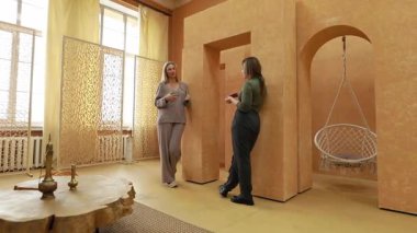 İki kadın şık bir şekilde tasarlanmış bir odada sohbet ediyorlar. Açık renkler, asılı bir sandalye ve dekoratif bölümler. Atmosfer sıcak ve davetkar, etkileşim teşvik edici..