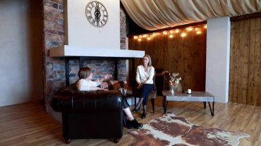 İki kadın sıcak, davetkar bir oturma odasında rahatça oturur ve kırsal dekorlar ve yumuşak ışıklandırmayla çevrili bir şekilde sohbet ederler. Atmosfer rahat ve rahat..