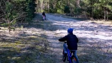 İki çocuk uzun ağaçlarla çevrili kumlu bir orman patikasında bisiklet sürmekten zevk alıyor. Baharda açık havayı keşfederken güneş ışıl ışıl parlıyor.