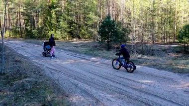 Sakin bir orman ortamında, bir çocuk kumlu bir patika boyunca bisiklet sürerken bir ebeveyn bebek arabasını itiyor. Güneş ağaçların arasından parlıyor, gezileri sırasında huzurlu bir atmosfer yaratıyor..