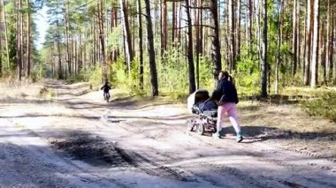 Bir anne, çocuğu yakınlarda bisiklet sürerken bir bebek arabasıyla huzurlu bir orman yolunda gezinir. Güneş ışığı ağaçların arasından süzülüp sakin bir atmosfer yaratır..