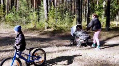 Bir çocuk, annesi yakınlarda bir bebek arabasını iterken kir içinde bisiklet sürüyor. Çevrede dallardan süzülen uzun ağaçlar ve güneş ışığı var..