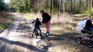 Bir anne, ağaçlarla çevrili toprak bir patika boyunca bisiklet sürme alıştırması yaparken çocuğuna rehberlik eder. Küçük bir köpek yakından izler, günün keyifli atmosferini artırır..