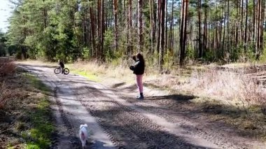 Genç bir bisikletçi, sakin bir orman ortamında doğanın güzelliğini yakalayan başka bir kişi ise kirli bir yolda ilerliyor. Güneş ışığı ağaçların arasından süzülüyor.