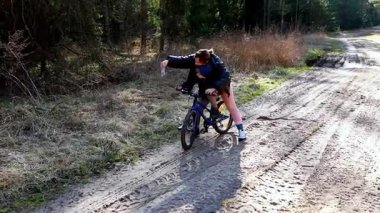 Bir çocuk ağaçlarla çevrili bir toprak yolda bisiklet sürme alıştırması yapıyor. Bir yetişkin, çocuğu koltuğun arkasını tutarak denge ve güveni teşvik ederek geçindirir..