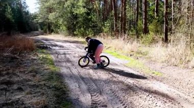 Güneşli bir bahar gününde uzun ağaçlarla çevrili kayalık bir orman patikasında bisiklet süren bir bisikletçi görülüyor. Çevre yeşil ve canlı, açık hava deneyimini geliştiriyor..