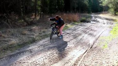Bir anne, ağaçlar ve doğa güzellikleriyle çevrili güneşli bir günde, ormandaki toprak bir patikada bisiklet sürmekten zevk alır..