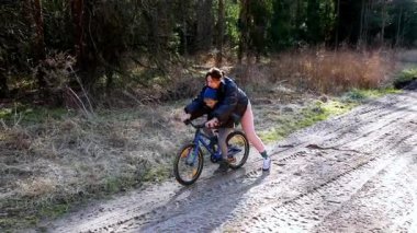 Bir çocuk gün ışığında güzel bir orman ortamında bir yetişkinin yardımıyla bisiklet sürme alıştırması yapıyor. Yol kumlu, yeşilliklerle çevrili..