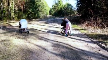 Bir kadın bebek arabasını iterken, diğeri sessiz bir orman yolunda bisiklet sürüyor. Güneş ağaçların arasından parlıyor, sakin bir atmosfer yaratıyor. Dışarıda bir gün için mükemmel..