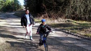 Bir kadın, yakınlarda bir bebek arabası park halindeyken ormanda kirli bir patikada bisiklet süren bir çocuğun yanında yürüyor. Hava aydınlık ve güzel. Açık hava aktiviteleri için mükemmel..
