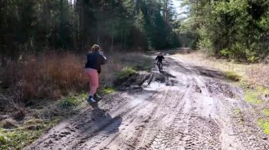 Bir bisikletçi çamurlu bir orman yolunda giderken bir fotoğrafçı yakınlardaki olayı yakalıyor. Güneş ışığı ağaçların arasından süzülüp açık hava atmosferini güçlendirir..