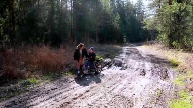 Bir kadın, bisikletini ağaçlarla çevrili bir patika boyunca sürerken genç bir çocuğu destekliyor. Güneşli öğleden sonra doğadaki ortak deneyimlerine sıcaklık katar..