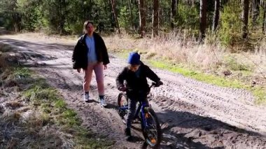 Genç bir çocuk, ailesi yakından izlerken ağaçlarla çevrili toprak bir patikada bisiklet sürme alıştırması yapıyor. Açık hava aktiviteleri için harika bir gün..