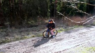 Bir anne, çocuğuyla birlikte yemyeşil ağaçlarla çevrili toprak bir yolda bisiklet sürüyor. Güneşli ormanlarda dolaşırken, doğanın tadını çıkarırlarken, keyifli bir anı paylaşıyorlar..
