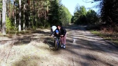 Bir kadın bisiklete binen bir çocuğa yardım ederken bir köpek de sakin bir orman ortamında onların yanında koşuyor. Canlı yeşil ağaçlar ve toprak patika mükemmel bir açık hava macera ortamı yaratır..
