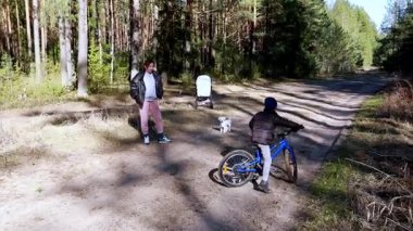 Bir çocuk mavi bir bisiklete binerken bir ebeveyn orman patikasının yakınlarını izliyor. Güneş ışığı ağaçların arasından süzülüp, sakin bir bahar atmosferi yaratır..