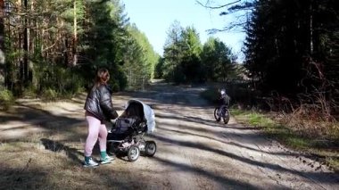 Bir anne, huzurlu bir orman ortamında bir bebek arabasını toprak bir patika boyunca iter. Küçük bir çocuk yakınlarda bisiklet sürüyor. Güneş ağaçların arasından parlıyor, sıcak bir atmosfer yaratıyor..