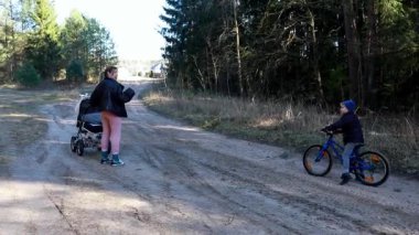 Bir kadın, küçük oğlu yakınlarda bisiklet sürerken bir bebek arabasını toprak yolda itiyor. Güneş ağaçların arasından parlıyor ve sıcak bir atmosfer yaratıyor. Doğada birlikte güzel vakit geçiriyorlar..