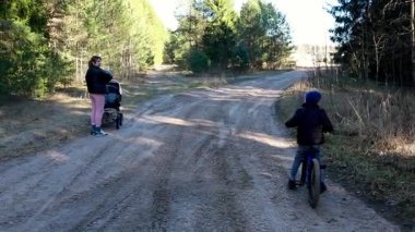 Bir anne, çocuğunun yanında bebekle gezer. Baharda yemyeşil bir ormanda, manzaralı bir toprak yolda bisiklete biner..