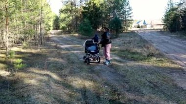 Bir anne çocuğunu ıssız bir orman yolunda bisiklete bindirirken bebek arabasını iter. Sahne huzurlu, uzun ağaçlar ve doğal yeşilliklerle çevrili..