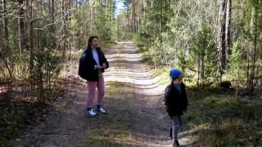 İki aile ferdi, uzun ağaçlar ve yeşilliklerle çevrili bir orman patikası boyunca canlı bir yürüyüşün tadını çıkarıyor. Huzurlu bir ortamda temiz hava ve güneş alıyor..