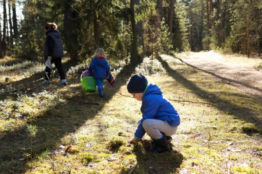 İki çocuk, biri yerde çömelmiş, diğeri kova taşıyor, yanında bir yetişkinle birlikte güneşli bir ormanda yürüyorlar. Atmosfer neşeli, güzelliği vurguluyor..