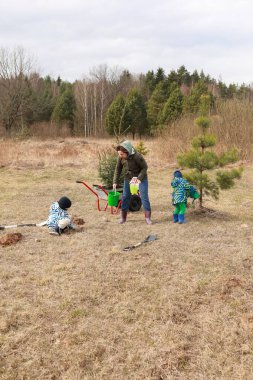 Bir aile kırsal alanda ağaç dikerek dışarıda zaman geçirir. Yetişkinler toprağı hazırlarken, çocuklar sulamaya katılıyorlar. Baharın büyümesini temsil eden yeşil ve huzurlu bir ortam..