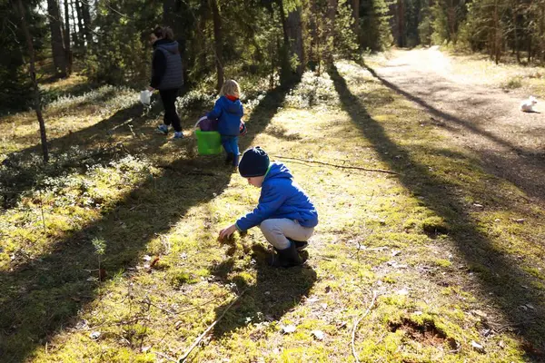 İki çocuk güneşli bir ormanda yiyecek ararken, biri yosunlu zeminde çömelmiş, diğeri kovada böğürtlen topluyor, dışarıda keyifli bir deneyim yaratıyor..