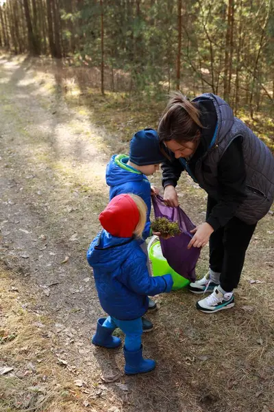 Ormanlık bir alanda güneşli bir günde, bir anne iki küçük çocuğuna bitki toplamada yardım eder. Çocuklar mutlu mesut nişanlılar, renkli ceketler ve şapkalar giyiyorlar..