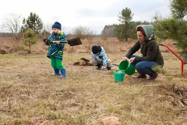 Bir kadın, açık bir çimenlikte genç ağaçlar dikerken iki çocuğa yardım ediyor. Çocuklar serin hava için sıcacık giyinirken kürek ve su kutuları kullanıyorlar..