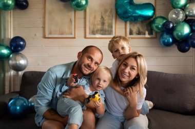 Dört kişilik bir aile renkli balonlarla çevrili bir kanepede toplanıyor. İki çocuk oyuncak arabalarla oynarken ebeveynler gülümsüyor, öğleden sonra evde keyifli bir anın tadını çıkarıyorlar..