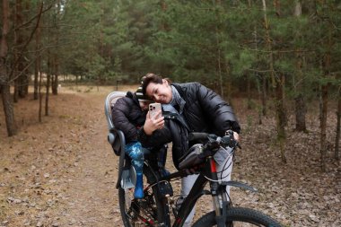 Bir ebeveyn ve çocuk ormanda bisiklet sürerken eğlenceli bir anı paylaşıyorlar. Çocuk kameraya gülümserken bisiklette bir koltukta oturur. Etrafları ağaçlar ve doğa ile çevrili..