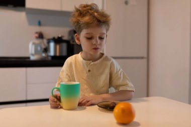 Genç bir çocuk mutfak masasında oturuyor, renkli bir bardaktan yudumlarken bir tabak kurabiye ve portakalı inceliyor. Güneş ışığı sıcacık alanı aydınlatır.