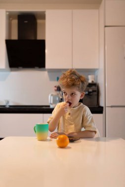 Mutfak masasında oturan genç bir çocuk renkli bir fincandan yudumlarken bir aperatif yiyor. Mutfakta modern cihazlar ve parlak bir öğleden sonrayı gösteren temiz bir düzen var..