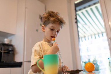 Genç bir çocuk elinde portakal parçalarıyla meyve yemeye odaklanmış. Mutfak iyi aydınlatılmış, turuncu ve renkli bardaklarla çevrili, neşeli bir atmosfer yaratıyor..