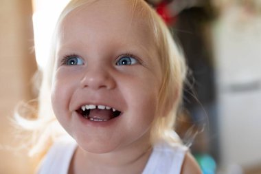 Küçük bir çocuk, büyük, bulaşıcı bir gülümseme sergiliyor sıcak, kapalı bir alanda. Parlak mavi gözler neşeyle parıldıyor, mutluluk ve masumiyet dolu bir anı yakalıyor..