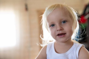 Kıvırcık açık sarı saçlı genç bir çocuk sıcak ve davetkar bir mekanda parlak bir şekilde gülümsüyor. Güneş ışığı yakındaki perdelerin arasından süzülerek neşeli atmosferi güçlendirir..