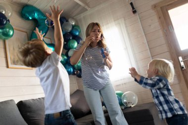 Çizgili tişörtlü bir kadın tarafından yaratılan baloncukları kovalayan iki genç çocuk. Renkli balonlar odayı süslüyor, neşeli bir atmosfer yaratıyor..