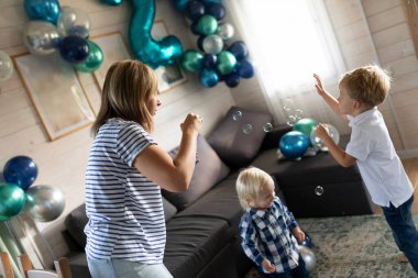Sıcak bir oturma odasında, anneleri yakınlarda dans ederken iki çocuk neşeyle kabarcıklarla oynuyorlar. Mekan, bir doğum günü kutlaması için şenlik balonları ve süslemelerle süslenmiş..