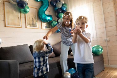 Bir kadın dekore edilmiş bir oturma odasında onlarla konuşurken çocuklar neşeyle kabarcıklarla oynuyorlar. Renkli balonlar şenlikli atmosfere ekleniyor.