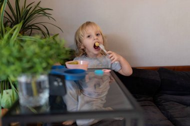 Küçük bir çocuk sıcak bir oturma odasında masada oturur ve mutlu bir şekilde atıştırmalık yer. Rahatlamış atmosfer, kapalı alandaki bitkiler ve yumuşak mobilyalarla tamamlanıyor..