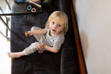 Açık gri giyinmiş genç bir çocuk, elinde küçük bir oyuncakla karanlık bir kanepeye rahatça oturur. Doğal ışık, sıcak oturma odasını doldurur ve sıcak bir atmosfer yaratır..