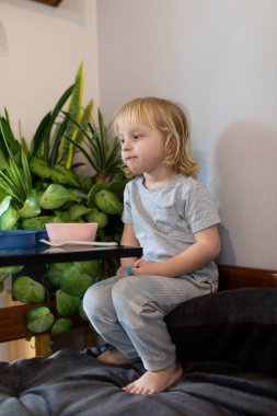 Genç bir çocuk düşünceli bir şekilde yemyeşil bitkilerle çevrili bir kanepede oturur, gündüz saatlerinde sakin ve düşünceli bir atmosfer sergiler..