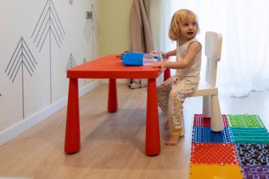 Küçük bir çocuk renkli paspaslar ve oyuncak kutularıyla süslenmiş bir odada kırmızı bir masada oturuyor. Güneş ışığı pencereden içeri akar ve yaratıcılık için neşeli bir atmosfer yaratır..